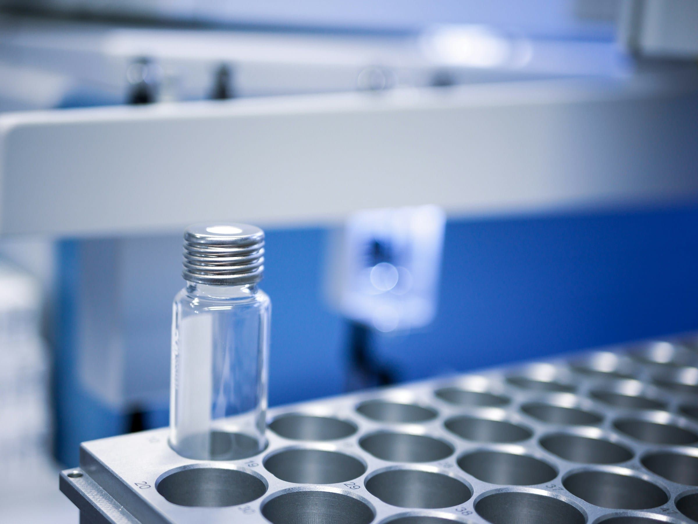 A single glass sample vial standing in a metal autosampler tray, lab equipment in cool blue light behind it